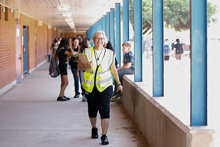 A woman in a yellow safety vest smiles as she walks through an outdoor corridor with students behind her