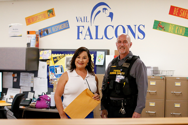 A woman in a white sleeveless shirt and a man in a school safety vest smile in the front office